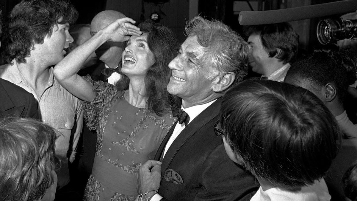 In this 1972 file photo Jacqueline Kennedy Onassis, center left, stands with conductor-composer Leonard Bernstein, center right, as she shields light from her eyes while looking up at the stage in the John F. Kennedy Center for the Performing Arts, in Washington, D.C. Bernstein would have turned 100 this year.