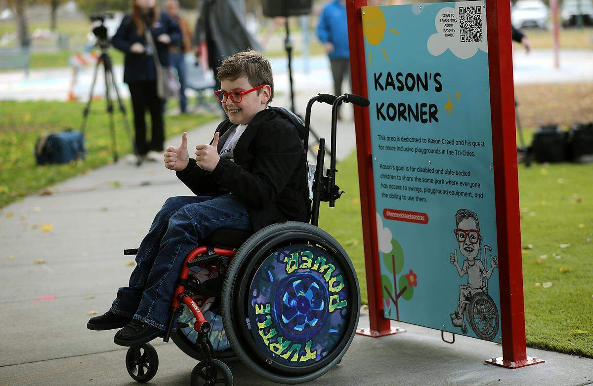 Kason Creed gives a thumbs up while family and friends take photos of him near the sign in his honor next to the new Kason’s Korner inclusive playground area at the Playground of Dreams in Kennewick’s Columbia Park.