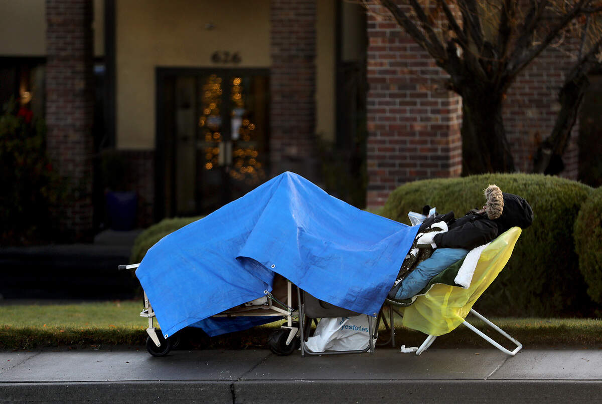A bundled up homeless person sleeps on the sidewalk in a folding chair partially under a tarp that also covers a wagon filled with  belongings on Columbia Center Boulevard on Dec. 23, 2024 in Kennewick.