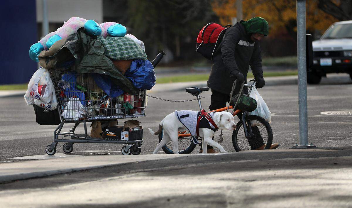 A homeless man pulling his belongings in a shopping cart while pushing a bike and  holding a leash to his dog crosses Edison Street at the West Clearwater Avenue intersection in Kennewick. on Nov. 25, 2024 
