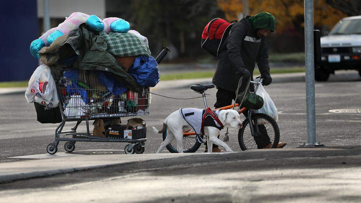 A homeless man pulling his belongings in a shopping cart while pushing a bike and holding a leash to his dog crosses Edison Street at the West Clearwater Avenue intersection in Kennewick. on Nov. 25, 2024
