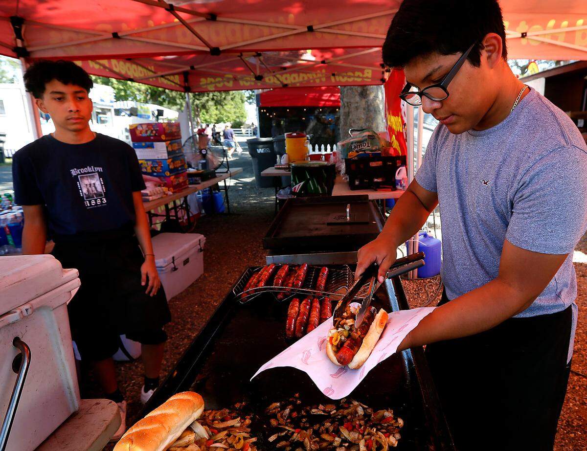 Juan Villa Deleon, left, and Joseph Marroquin work together to serve up a bacon wrapped hot dog for a customer at their booth at the Benton Franklin Fair & Rodeo in Kennewick.