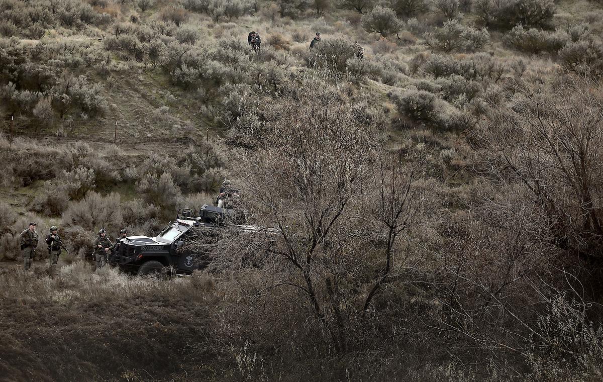Members of the Tri-Cities Regional SWAT team stand behind their armored vehicle while a drone hovers above a clump of trees in a ravine searching for a Kennewick murder suspect.