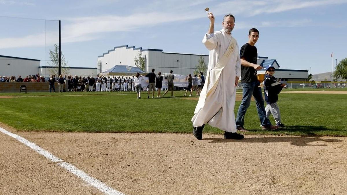 Deacon Bill Mick, left, along with graduate Isaac Nelson and his brother Luke, 6, bless first base Saturday during the dedication of Tri-Cities Prep new softball and baseball field at the school in Pasco. The Ray Whitlow Memorial Athletic Complex was named in the memory of the school’s first athletic director.