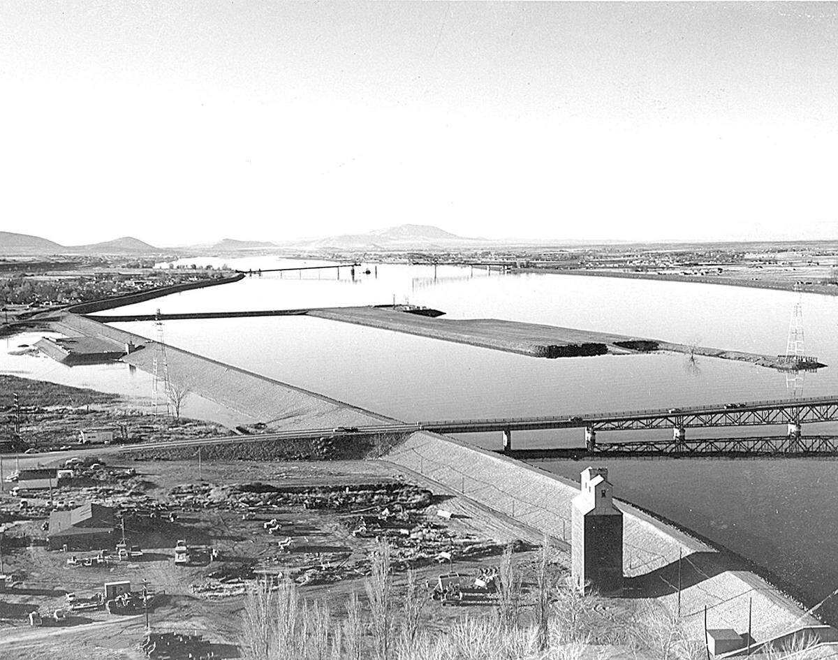Thirty undeveloped acres next to the blue bridge in the distance is one of the region’s hottest development prospects. This historic photo shows the Kennewick shoreline some time after the McNary Pool formed behind the dam. This photo shows the early development of Clover Island, formed when  approximately 17 acres of fill material was piled nearly 20 feet deep on the high point of the submerged island. Pioneer Memorial bridge, completed in 1954 and now called the blue bridge, is shown nearly finished in the background.
