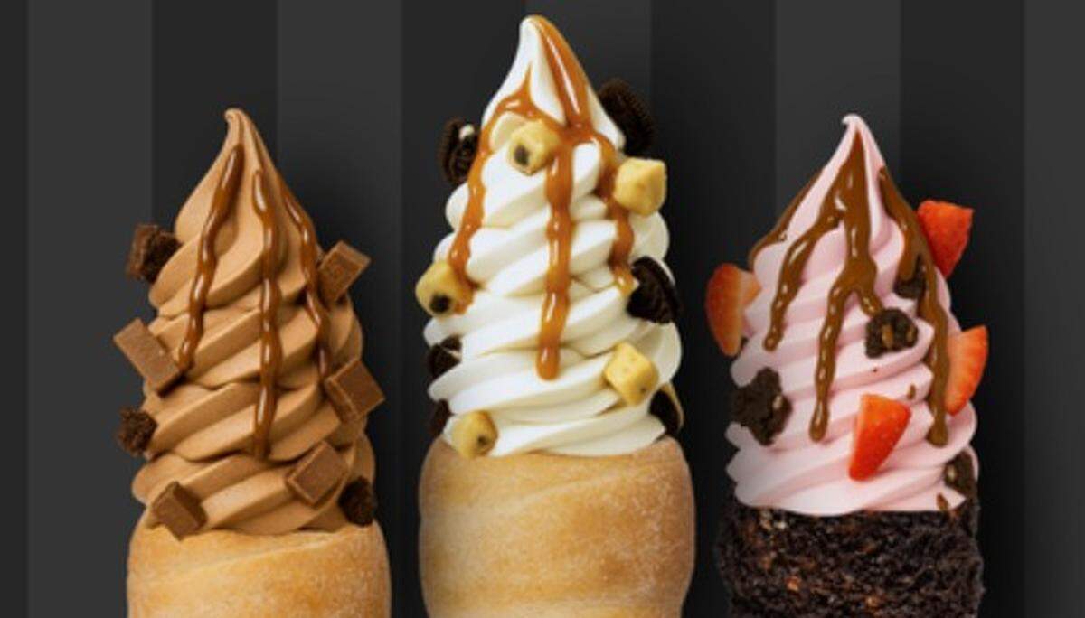 Three large, swirled soft-serve ice cream treats stand side-by-side against a dark, striped background. The three flavors are chocolate, vanilla, and strawberry, all topped with caramel drizzle and various toppings like chocolate chunks, hazelnuts, and fresh strawberries.