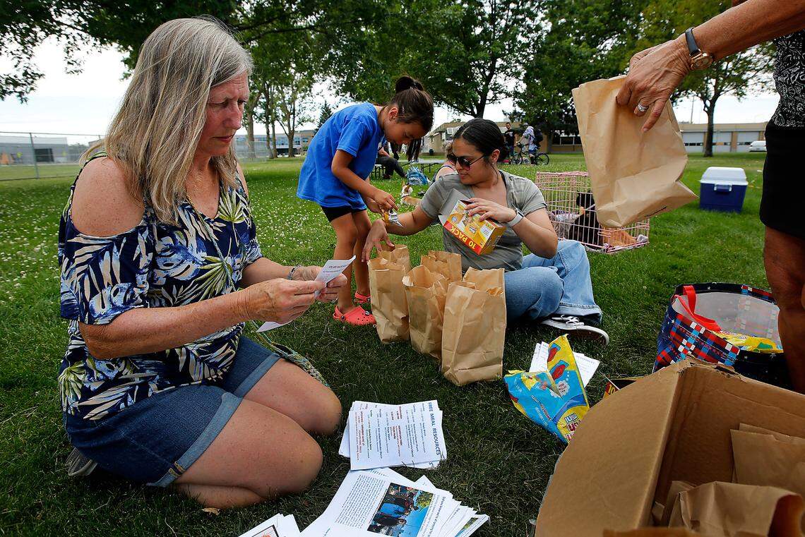 Volunteers Cindy Fish, six-year old Anaiyah Escamilla and her mother, Becky, from left, fill paper bags with toiletries, snacks and a flier about resources for homeless during a weekly free meal distribution on June 7, 2023 at Keewaydin Park in downtown Kennewick.