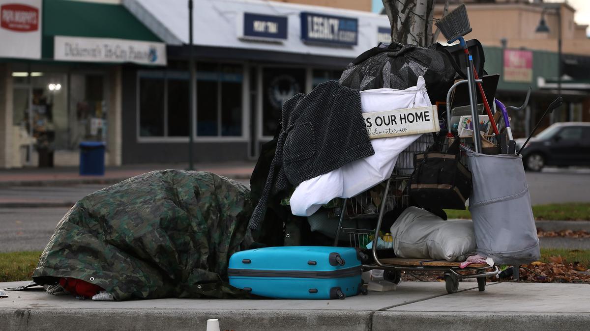 Homeless sleep under a tarp on the sidewalk near the Uptown Shopping Center in Richland near a cart filled with belongings and adorned with a sign saying, “Bless our home & all who enter.”