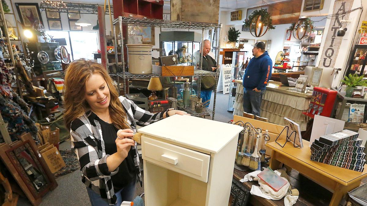 Amy Bergan works on refinishing a nightstand, while her husband, Jason, talks with customers Wednesday in their antique store, Bergan’s Timeless Treasures, in downtown Kennewick.