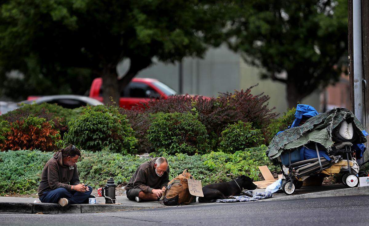 A pair of men and their dog sit on the sidewalk near wagons filled with their belongings on North Union Street near the WinCo grocery store off West Clearwater Avenue in Kennewick in August 2024.