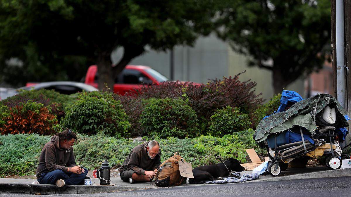 A pair of men and their dog sit on the sidewalk near wagons filled with their belongings on North Union Street near the WinCo grocery store off West Clearwater Avenue in Kennewick in August 2024.