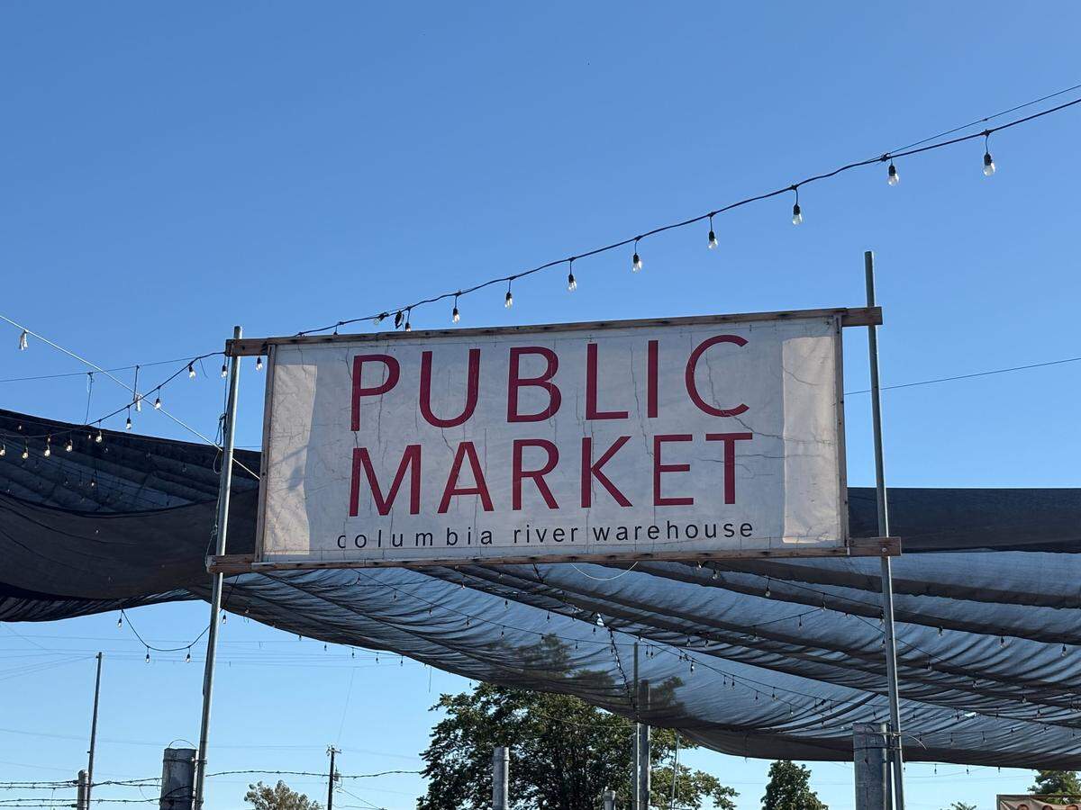 The entrance to the Public Market in Kennewick is marked by a large sign. An outside area sits behind the sign, with a covered lawn with children’s toys, a giant chess board, music playing and places to sit.