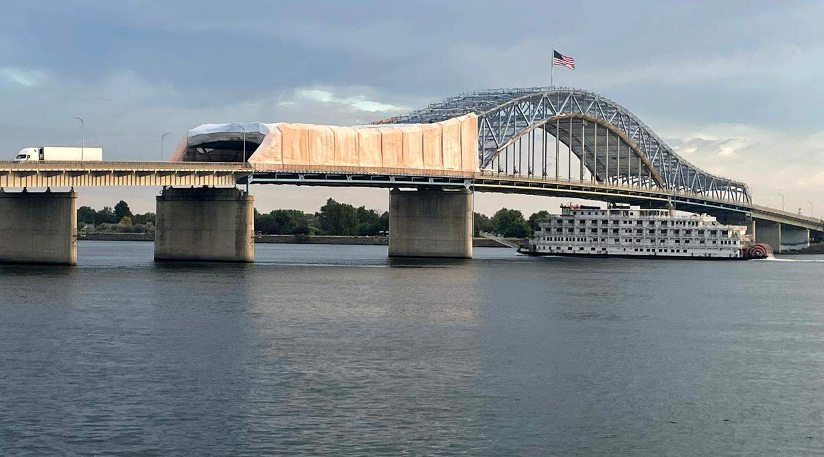 A “cocoon” of tarps on the south side of the span as a strong breeze unfurls the U.S. flag and a river paddle boat navigates up the Columbia River before workers started temporarily removing them Tuesday morning because of a wind advisory for the Lower Columbia Basin. The tarps are being used prevent debris falling into the river or onto passing vehicles during a year-long, $33.5 million paint project.