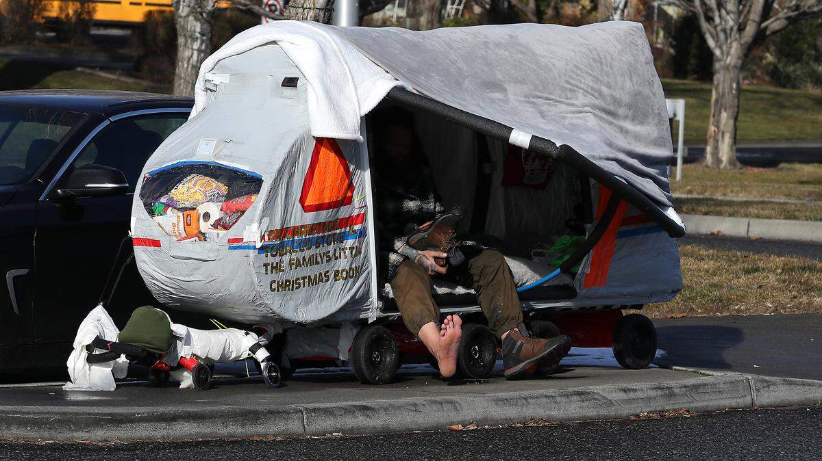 Homeless man in homemade wagon tent puts on his boots while sitting on the sidewalk in the Uptown Shopping Center in Richland on Feb. 5, 2025.