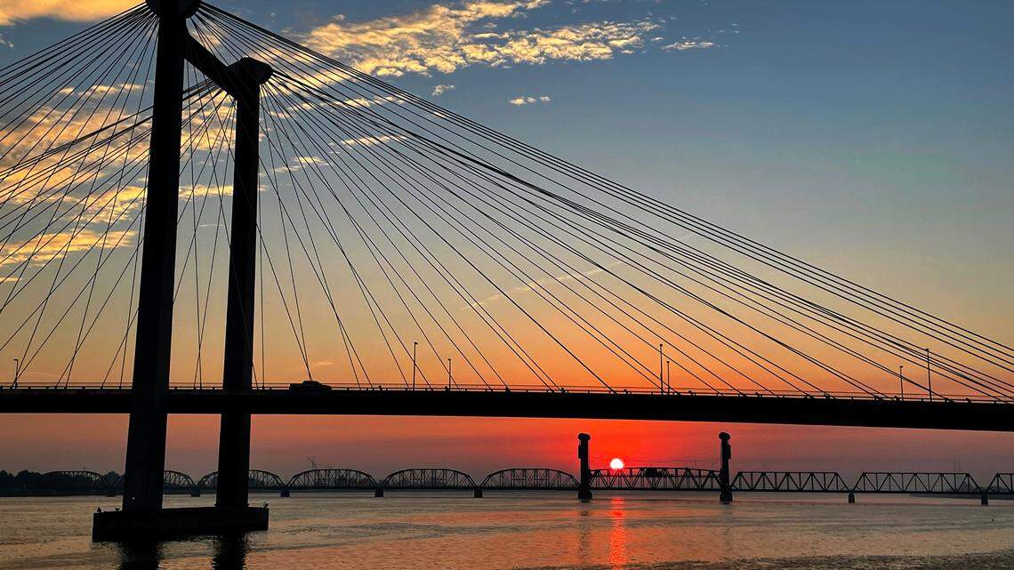 The sun rises over the Columbia River behind the cable bridge and railroad bridge between the Kennewick and Pasco shorelines.