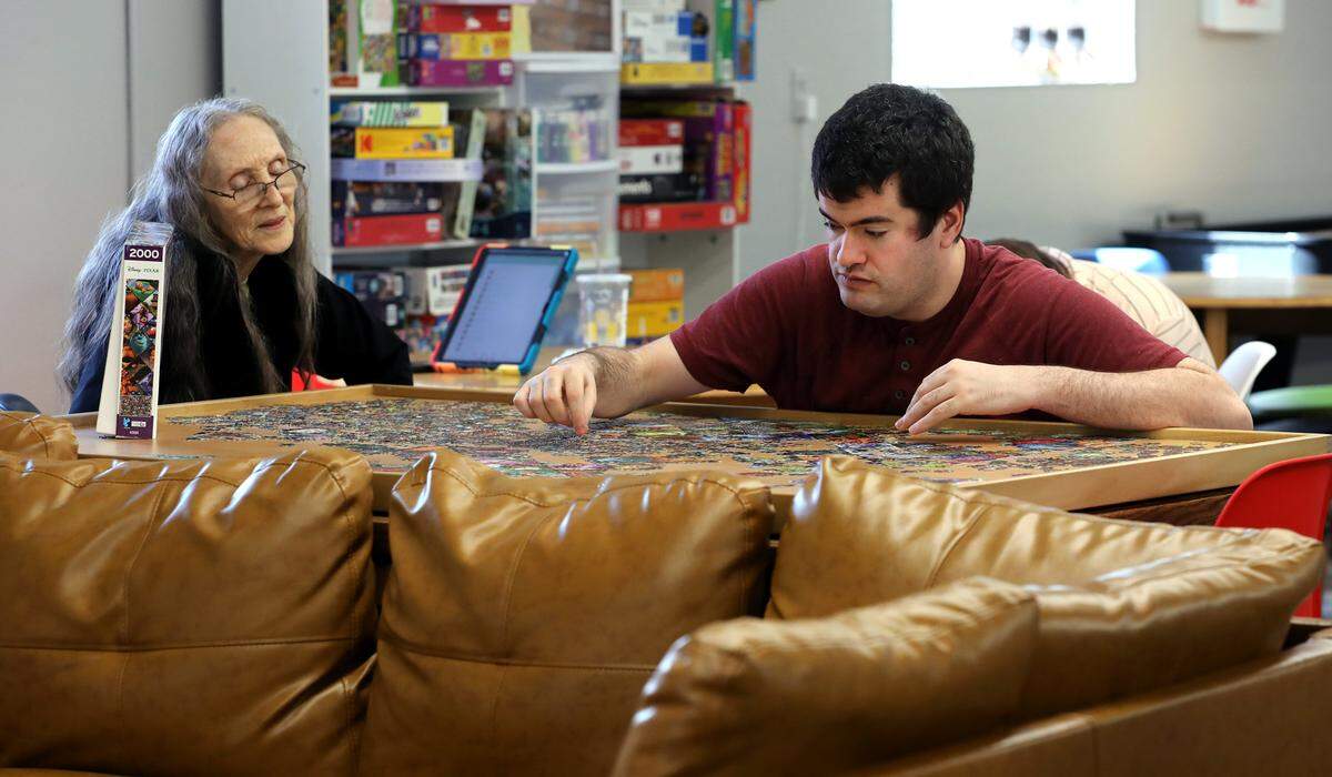 Community Center aide Kathy Kelley, left, sits with client Ian Hargan, 33, of Pasco, while he works on a large table top puzzle recently at the Columbia Ability Alliance facility in downtown Kennewick.
