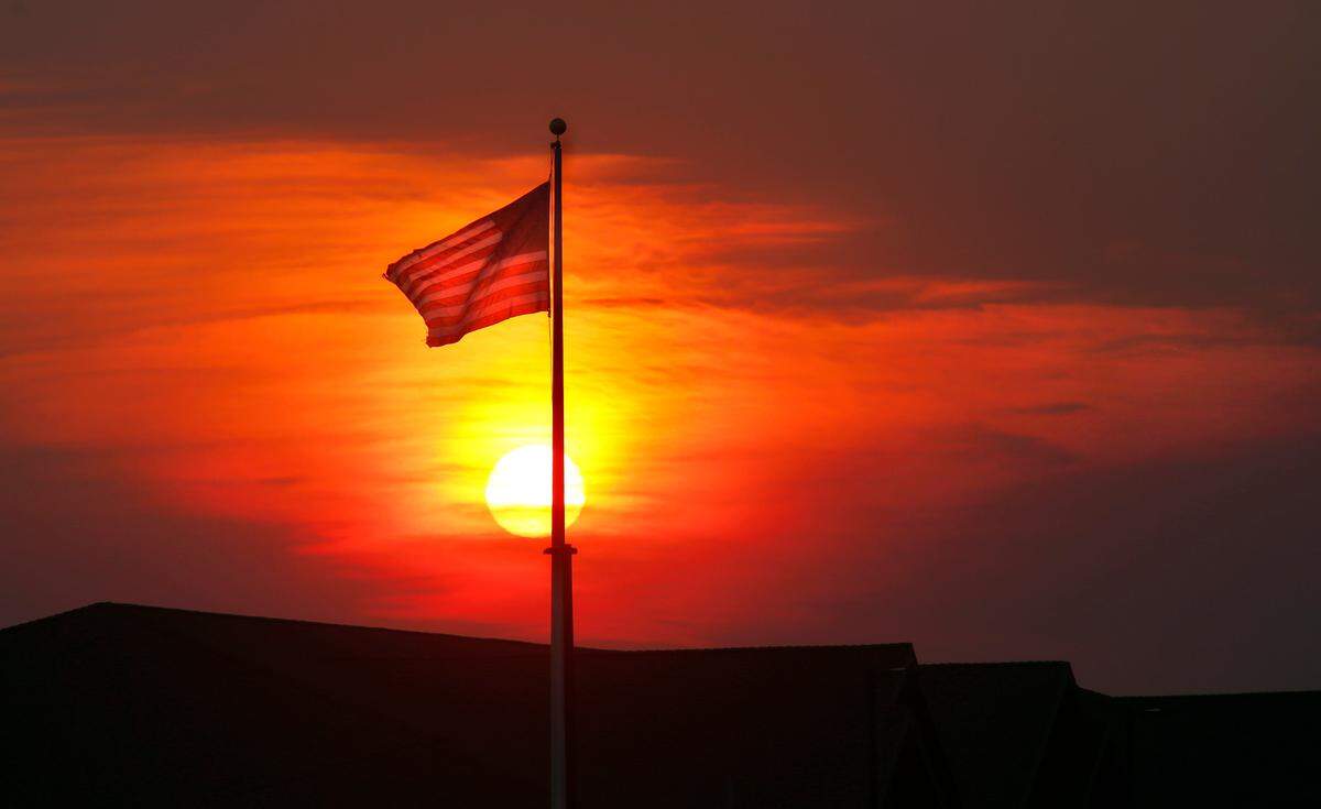 A hazy smoke-filled sky from wildfires in the region glows with brilliant hues of orange, yellow and red Wednesday morning as the sun rises behind a U.S. Flag in south central Kennewick.