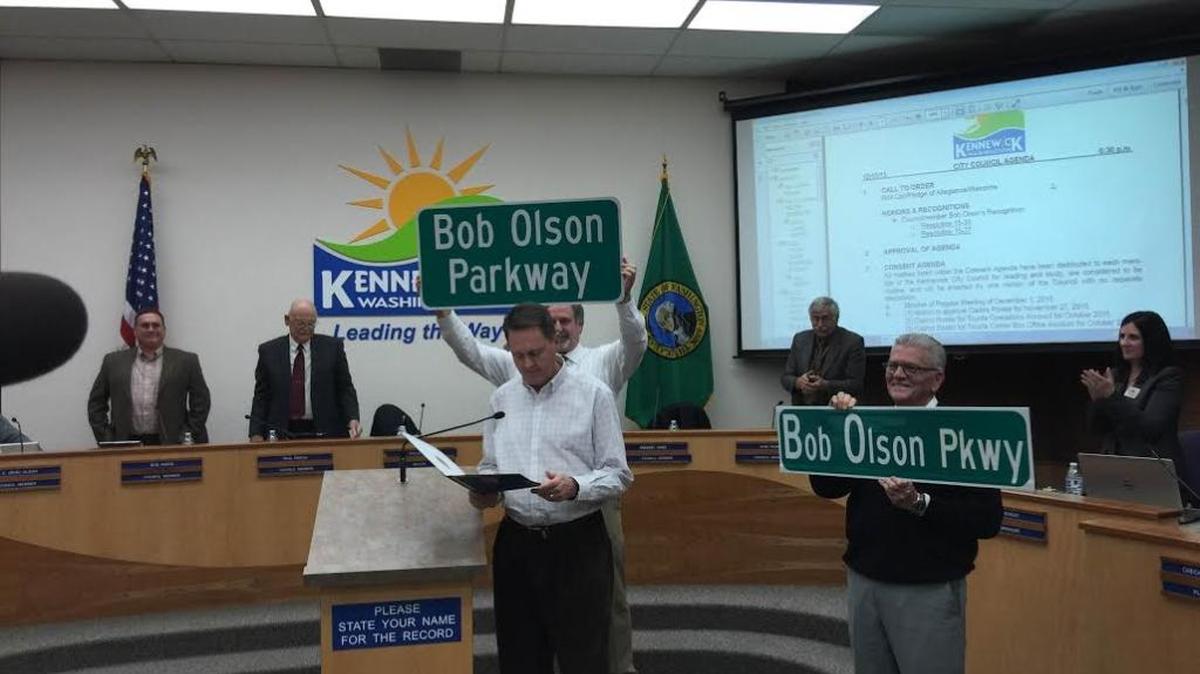 Kennewick Mayor Steve Young, right, and Public Works Director Cary Roe hold up signs that will be placed on a new road named for outgoing Councilman Bob Olson, while Councilman Greg Jones reads a proclamation in Olson’s honor.